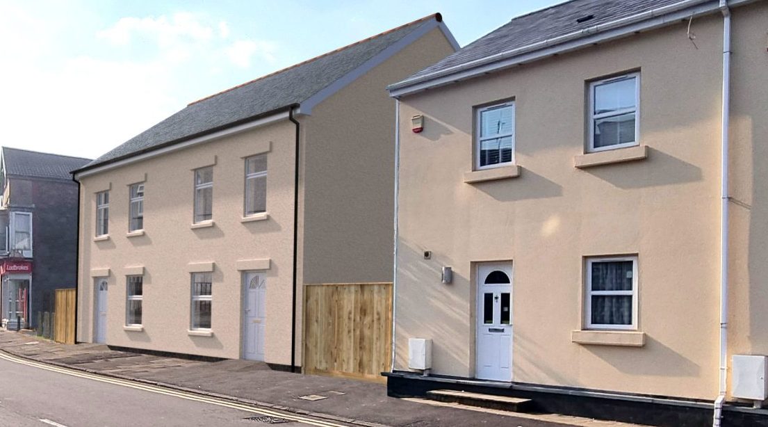 Trivallis Housing Landlord Wales A man in white walks past new light-colored townhouses on a sunny street with cars parked along the curb, with other residential buildings and a clear blue sky in the background. Safety checks keep these homes safe from hazards.