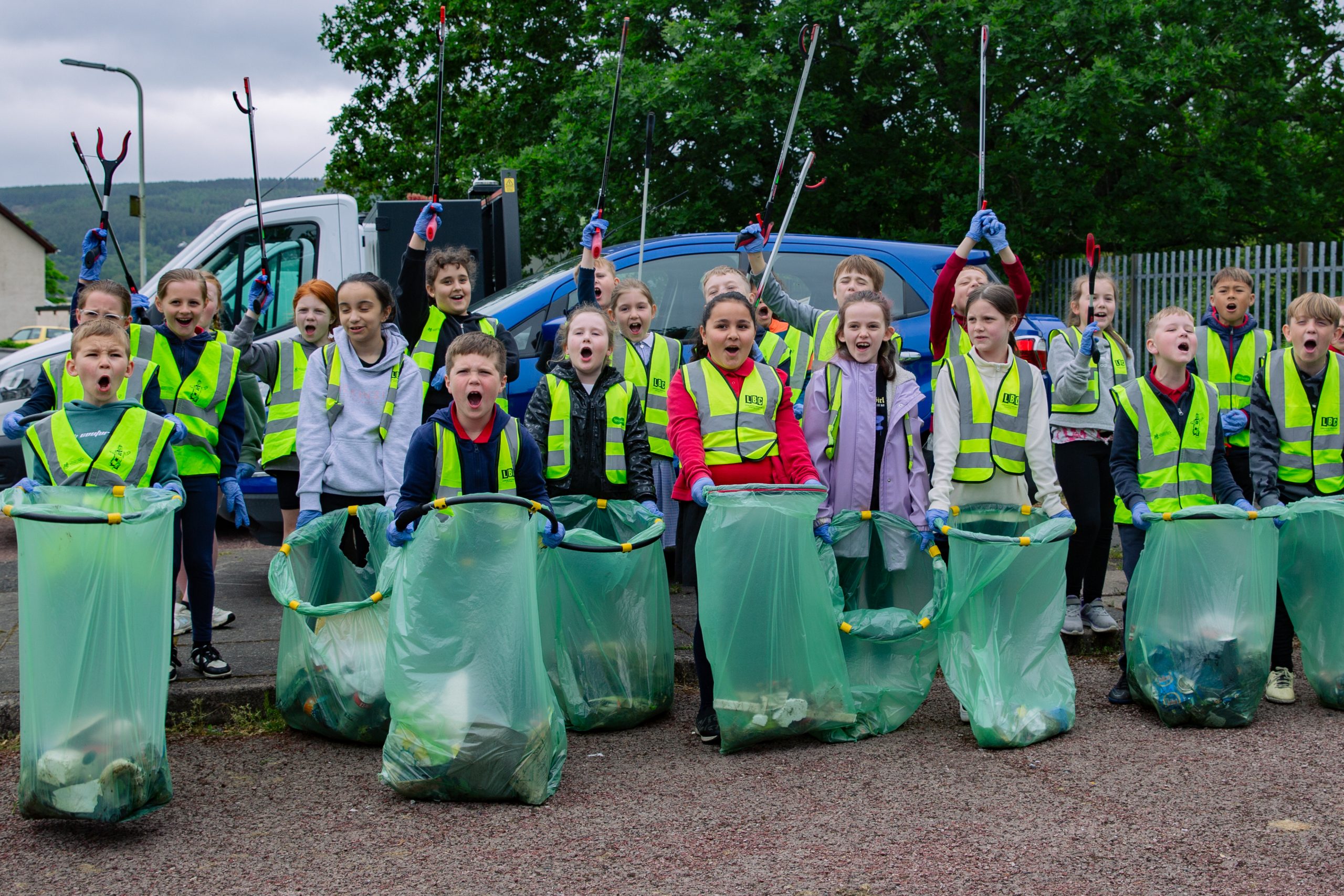 Trivallis Housing Landlord Wales A group of children in high-visibility vests stand in front of collected garbage bags, holding trash grabbers above their heads. They appear to be participating in a community cleanup event. There is a tree and some parked vehicles in the background.