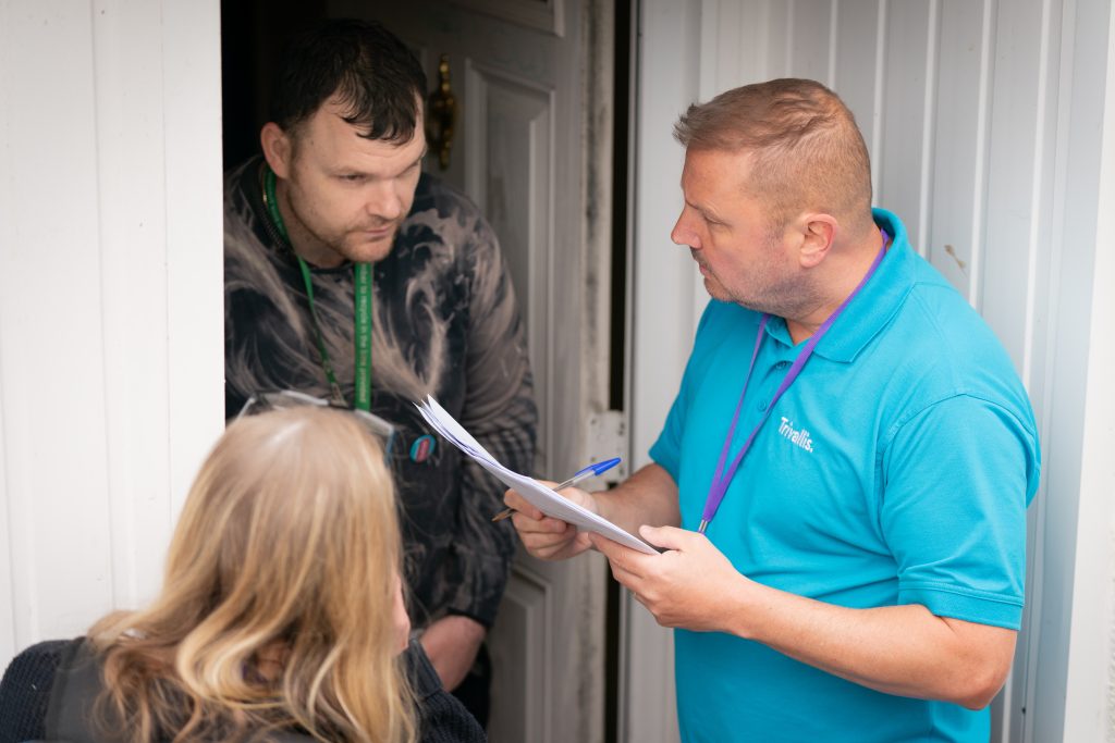 Trivallis Housing Landlord Wales A man in a blue shirt holding papers and a pen speaks to another man standing in a doorway. A woman with blonde hair stands in front of them, facing away from the camera. The interaction appears to be taking place at the entrance of a building.