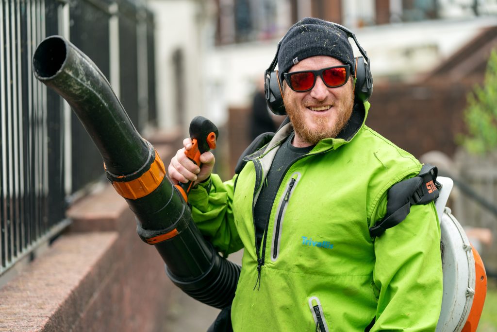 Trivallis Housing Landlord Wales A person smiles at the camera while holding a leaf blower. They are wearing a green jacket, red-tinted glasses, ear protection, and a black beanie. A railing and a building are visible in the background.