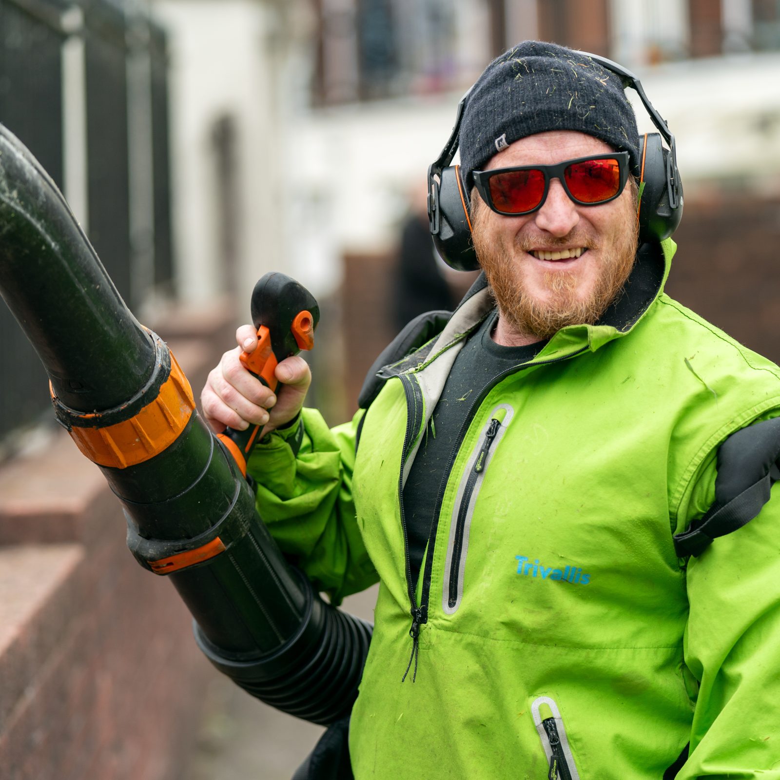 Trivallis Housing Landlord Wales A person smiles at the camera while holding a leaf blower. They are wearing a green jacket, red-tinted glasses, ear protection, and a black beanie. A railing and a building are visible in the background.