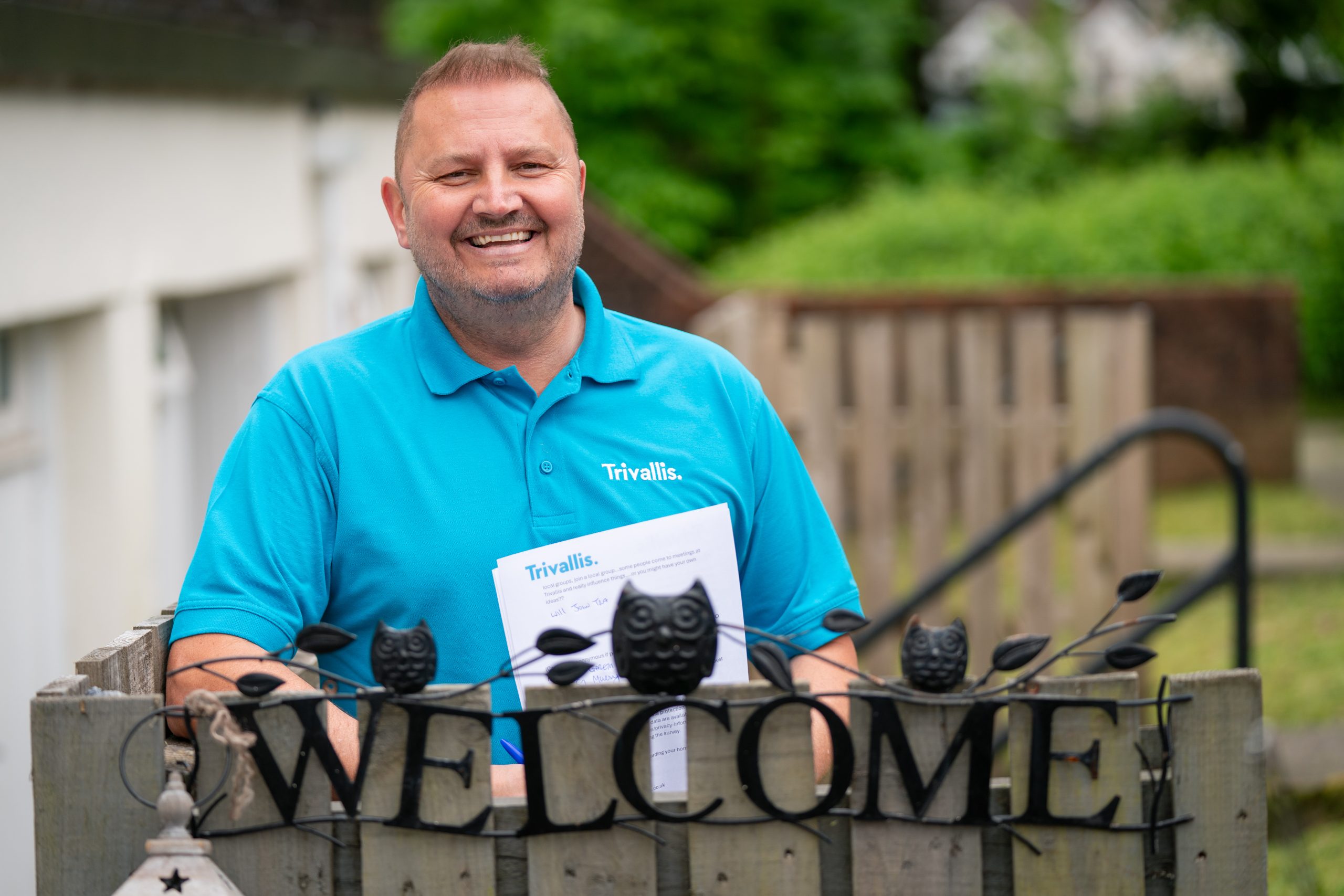 Trivallis Housing Landlord Wales A person in a blue shirt, holding a document, stands behind a decorative "WELCOME" sign featuring black owl figures. The person is smiling and appears to be in an outdoor setting with greenery and buildings in the background. The shirt has "Trivallis" printed on it.