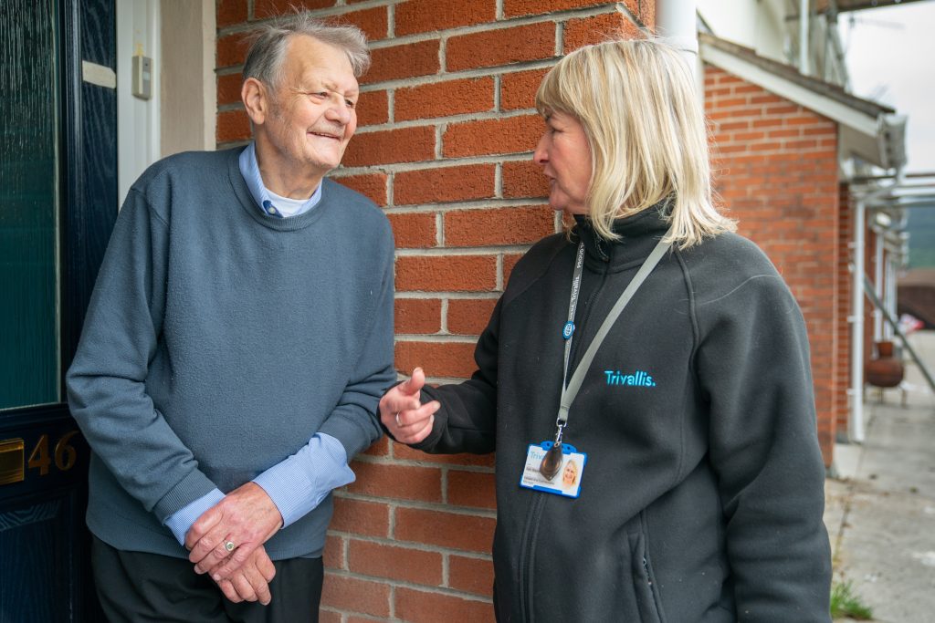 Trivallis Housing Landlord Wales An elderly man in a blue sweater stands at the door of house number 46, talking to a woman in a black jacket with a "Trivallis" logo. The woman wears a blue lanyard and ID badge. They appear to be engaged in conversation outside the brick house.