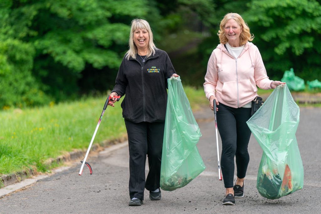 Trivallis Housing Landlord Wales Two women are seen walking on a paved path while participating in a litter-picking activity. They are carrying large green garbage bags and using grabber tools to pick up trash. The background is filled with greenery. Both are smiling.