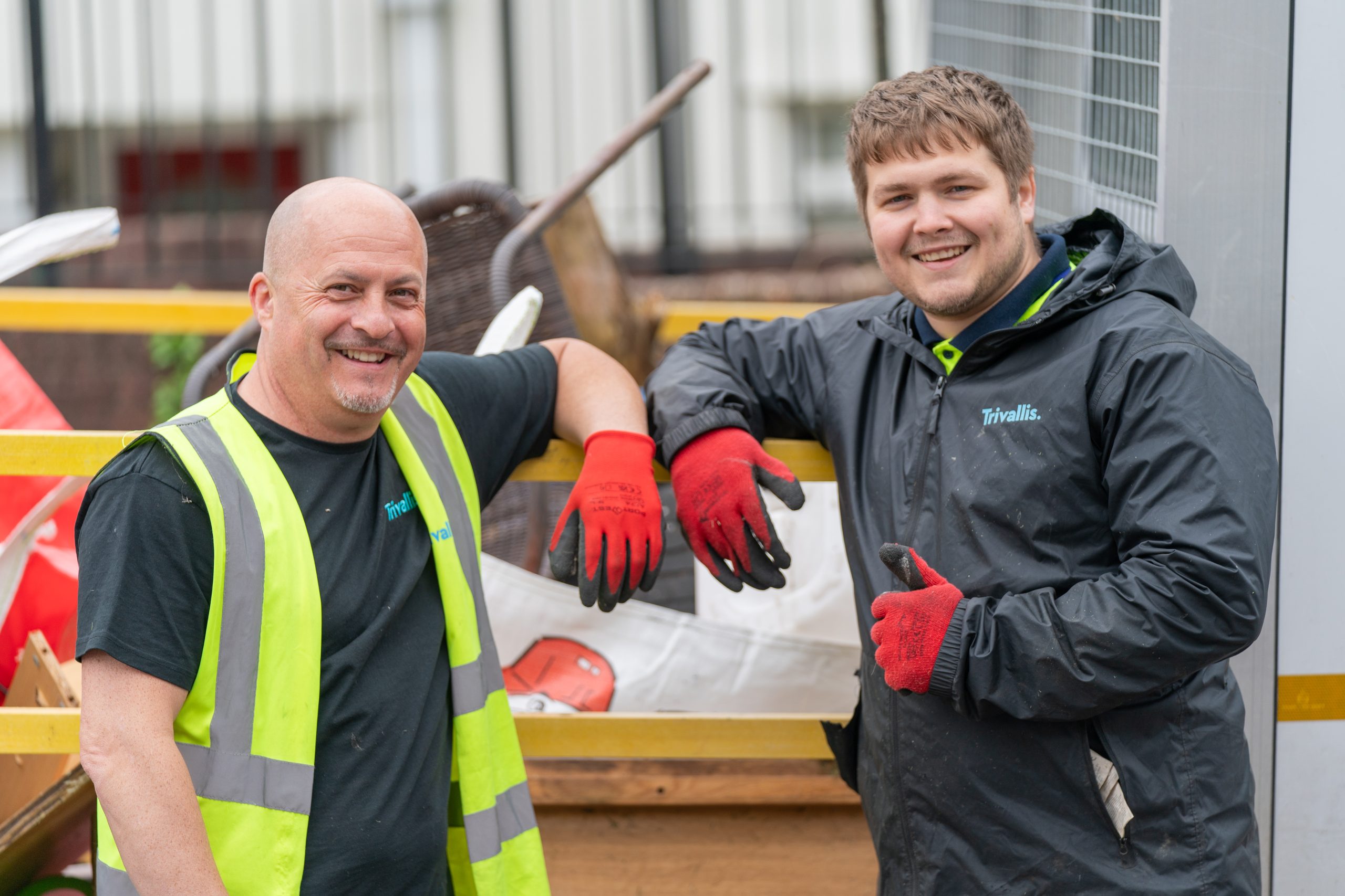 Trivallis Housing Landlord Wales Two men in work attire, one wearing a high-visibility vest and the other in a black waterproof jacket, both with red work gloves, are standing in front of a construction site. They are leaning on a railing and smiling at the camera.