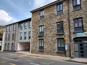 Trivallis Housing Landlord Wales A street view of a building with a stone façade and multiple windows. Adjacent to it is a newer building with a combination of stone and modern materials, featuring a small carport. A bus stop sign is visible on the sidewalk. The sky is partly cloudy.