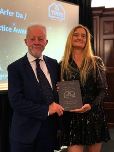 Trivallis Housing Landlord Wales A man and a woman stand together indoors, both wearing formal attire and smiling. The man holds a plaque that reads "TPAS / Good Practice Award 2023." There is a presentation screen in the background displaying the TPAS logo and part of the text "Good Practice Award.