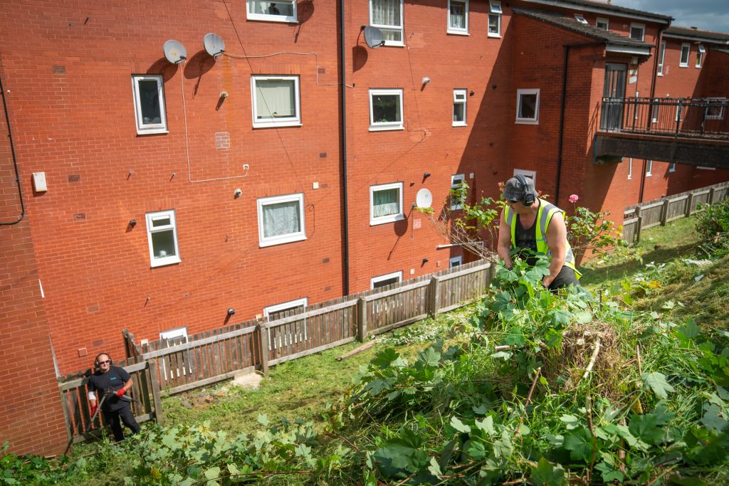 Trivallis Housing Landlord Wales Two people are working in a garden at the side of a brick apartment building. One person, wearing a black mask and bright yellow vest, is using a strimmer on greenery. The second person, wearing a dark shirt, is further back holding some garden tools.