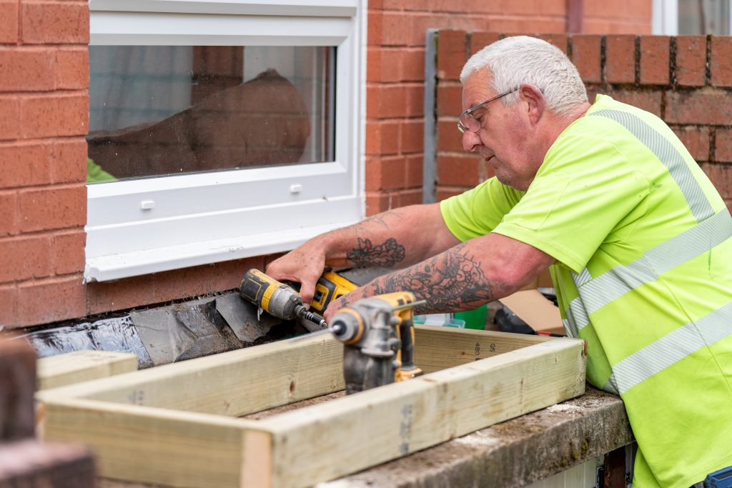 Trivallis Housing Landlord Wales A man wearing a bright green safety shirt and glasses uses a power drill to work on an outdoor construction project. He is assembling a wooden frame near a brick wall with a window, and has a tattoo on his right forearm.