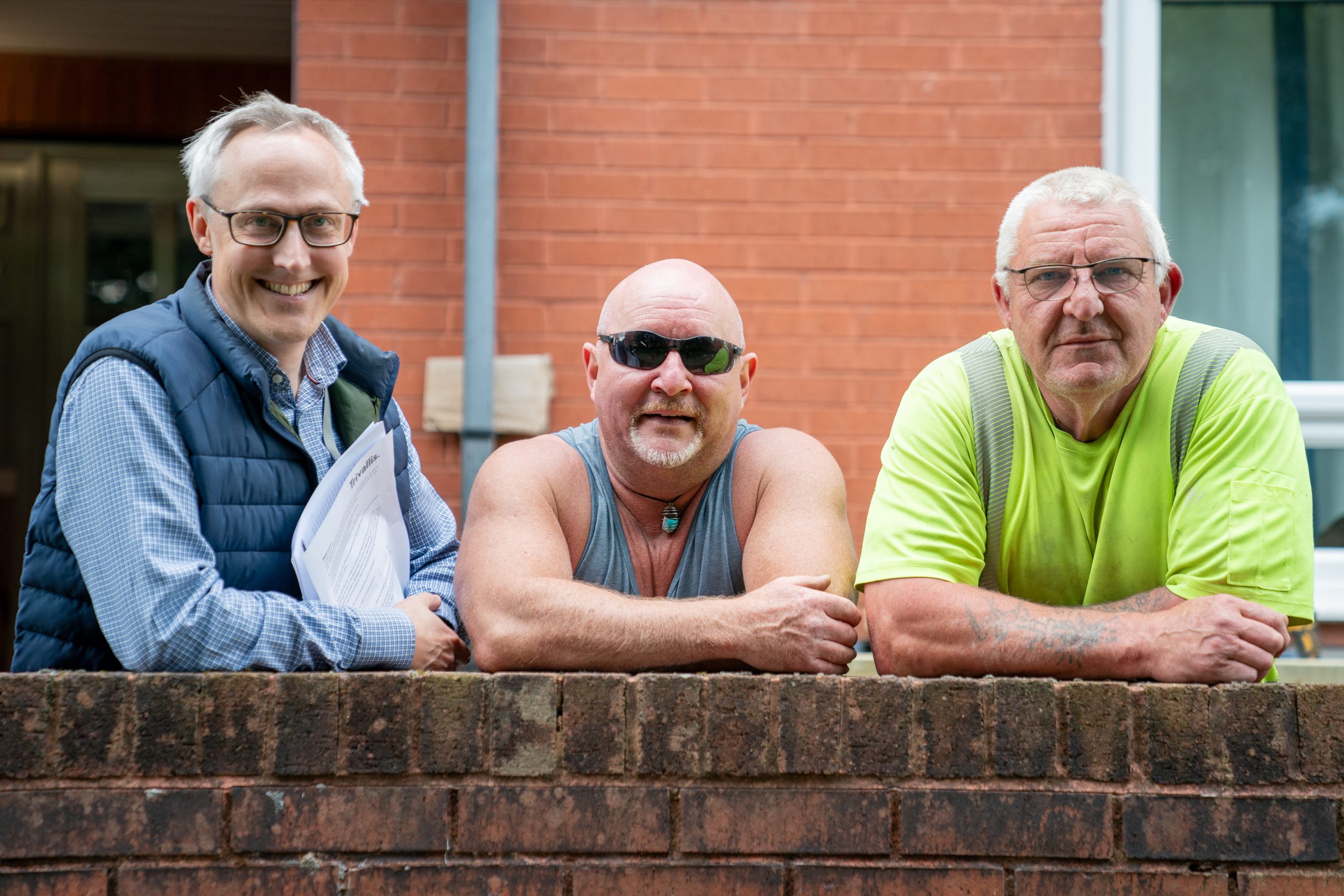 Trivallis Housing Landlord Wales Three men are standing and leaning on a brick wall. The man on the left is wearing glasses, a blue shirt, and a dark vest, holding papers. The middle man has a shaved head, sunglasses, and a gray tank top. The man on the right is wearing glasses and a neon yellow shirt.