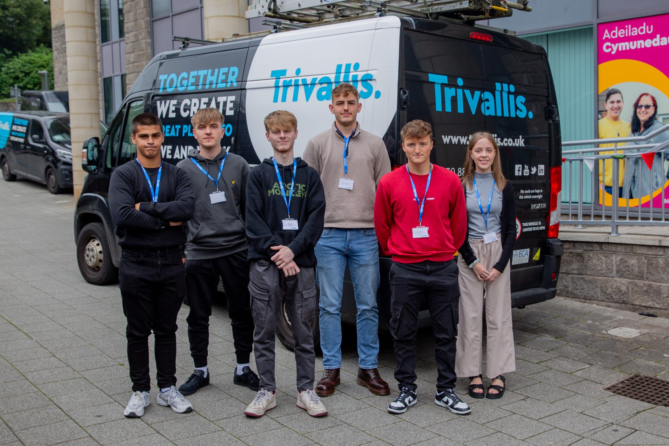 Trivallis Housing Landlord Wales Six apprentices standing in a line on a sidewalk, posing for a group photo. They are all wearing lanyards with nametags. Behind them is a black van with "Trivallis" branding and some text. A colorful advertisement is visible in the background.