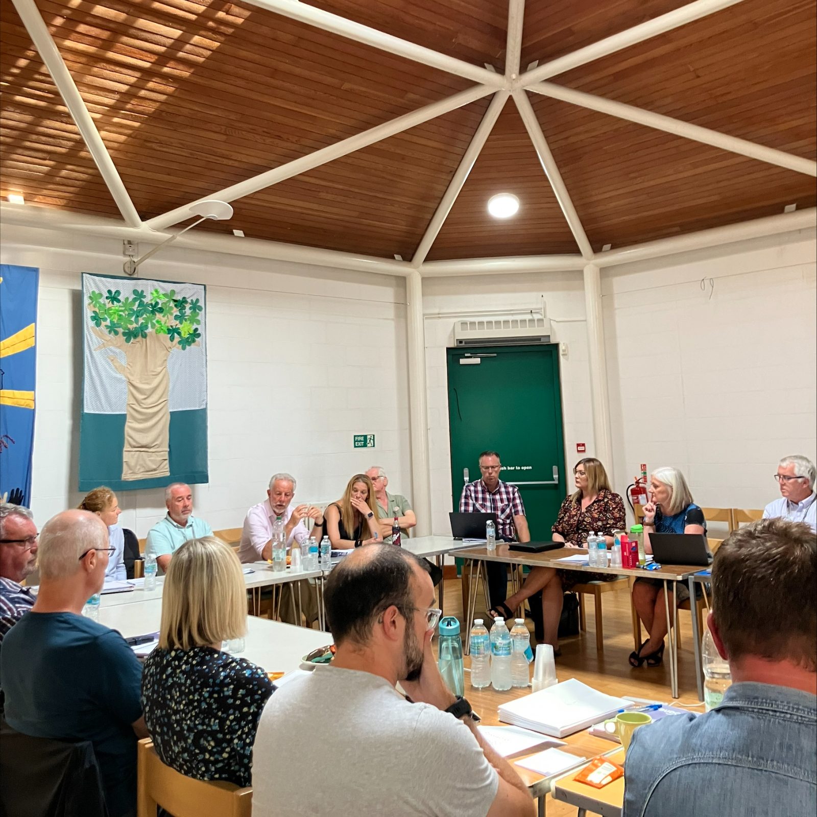 Trivallis Housing Landlord Wales A group of people sit around tables in a well-lit room with a wood-paneled ceiling. Some are talking while others listen. Various drinks and papers are on the tables. In the background, a green door and two wall hangings can be seen.