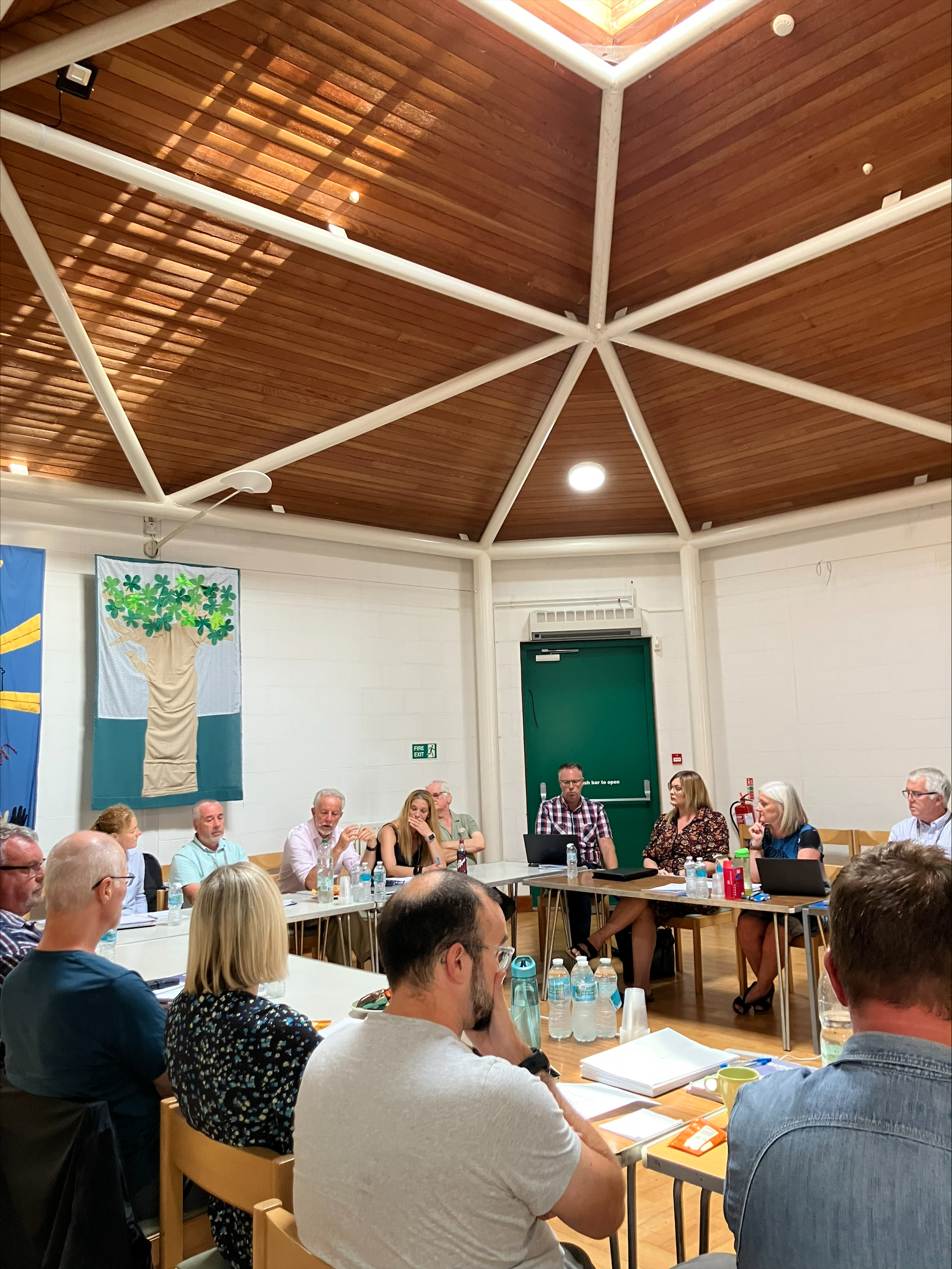 Trivallis Housing Landlord Wales A group of people sit around tables in a well-lit room with a wood-paneled ceiling. Some are talking while others listen. Various drinks and papers are on the tables. In the background, a green door and two wall hangings can be seen.