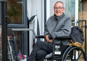 Trivallis Housing Landlord Wales A man in a gray sweater sits in a wheelchair outside a building with glass doors. He has short hair and wears glasses. His left hand rests on his lap, while his right elbow rests on a chair draped with a jacket next to him.