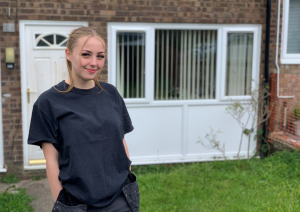 Trivallis Housing Landlord Wales A young woman with blonde hair is standing in front of a brick house with white-framed windows. She is wearing a dark t-shirt and has her hands in her pockets. The grassy yard is visible in the foreground.