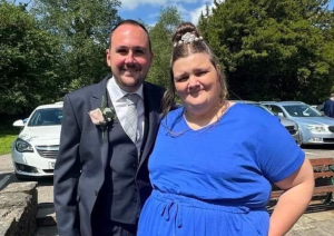 Trivallis Housing Landlord Wales A man and a woman are standing outdoors, smiling at the camera. The man is wearing a dark suit with a light-colored tie and a boutonniere, while the woman is dressed in a blue dress. There are several cars and trees in the background.