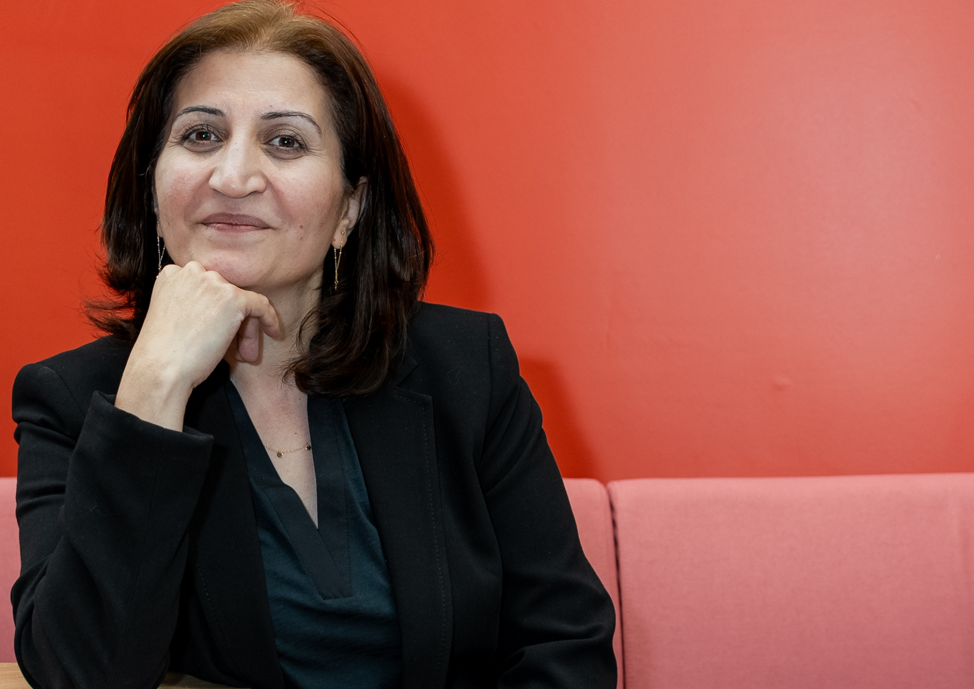 Trivallis Housing Landlord Wales A woman with shoulder-length dark hair and a dark blazer sits on a pink couch. She is smiling and resting her chin on her hand. The background is a bright red wall.