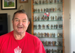 Trivallis Housing Landlord Wales A man in a red shirt stands in front of a glass display case filled with various action figures. The display case has several shelves, each densely packed with the figures. The background includes framed pictures and a part of a television.