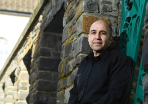 Trivallis Housing Landlord Wales A man in a black shirt leans against a stone wall outdoors, with a partially visible building in the background. He appears to be looking at the camera. There are green metal decorative elements attached to the wall.