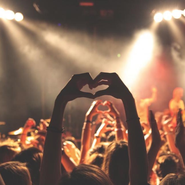 Trivallis Housing Landlord Wales A crowd at a concert with hands raised, some forming heart shapes. The scene is lit by overhead spotlights, creating a bright atmosphere among the audience and stage. The background is slightly blurred, focusing on the energetic crowd.