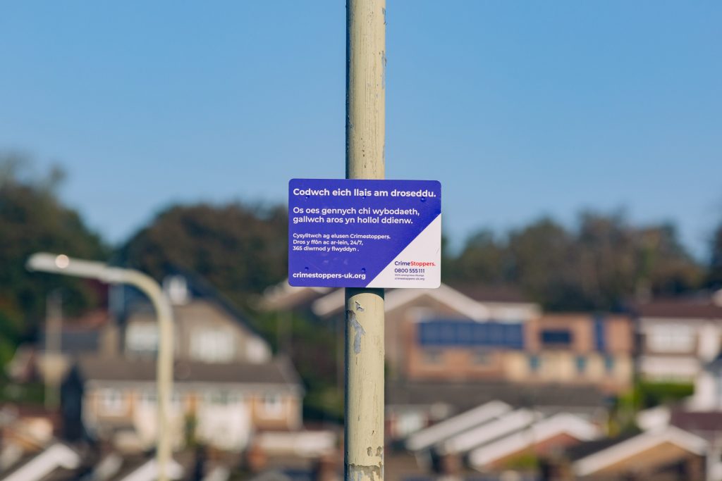 Trivallis Housing Landlord Wales A sign on a pole in front of a suburban neighborhood. The sign is in Welsh and English, encouraging people to report information about crime anonymously. Houses and trees fill the background.