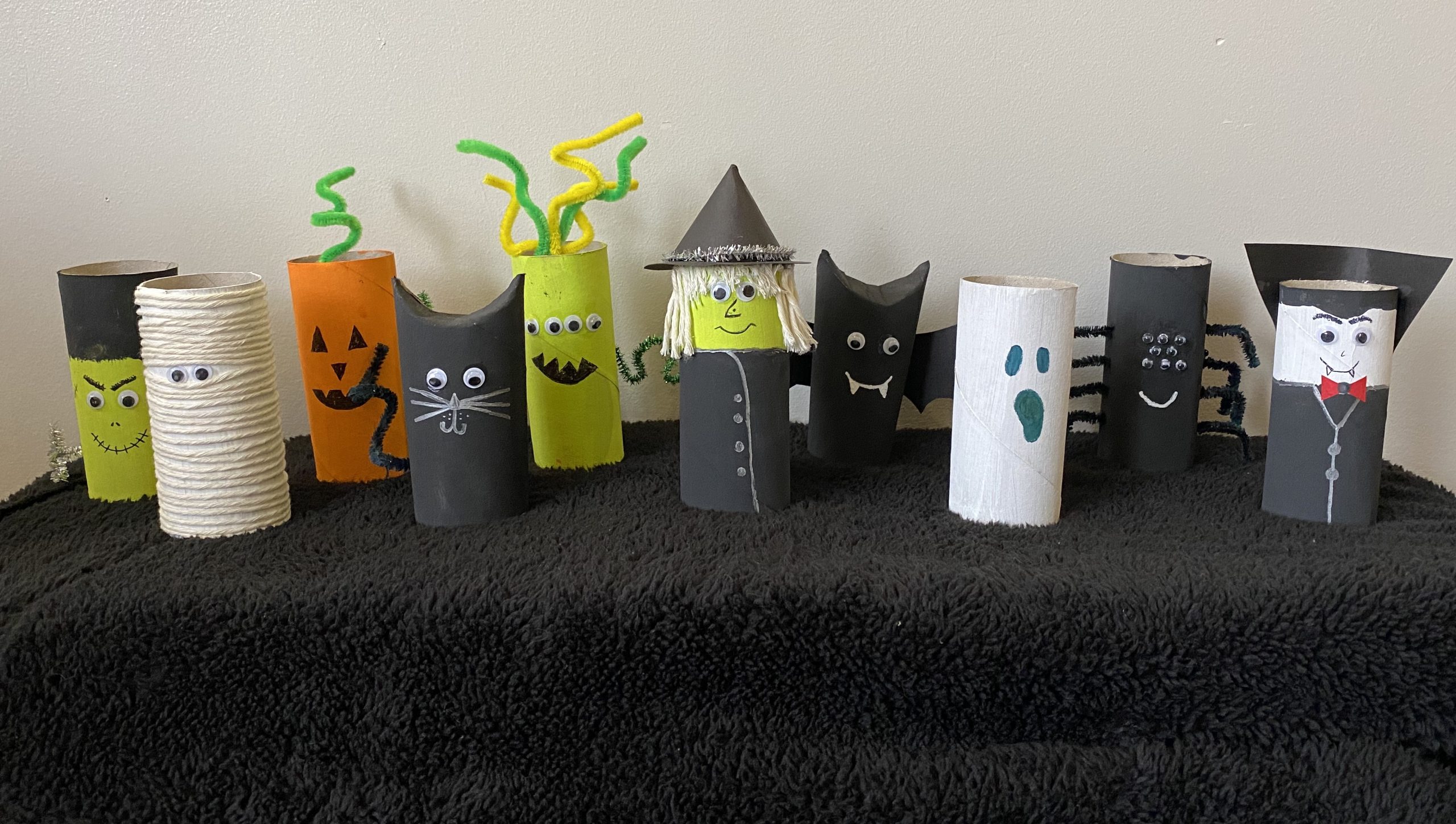 Trivallis Housing Landlord Wales A collection of Halloween-themed crafts made from toilet paper rolls, featuring characters like a witch, a ghost, a spider, a vampire, and others, each decorated with various materials like paint, pipe cleaners, and felt, displayed on a black cloth.