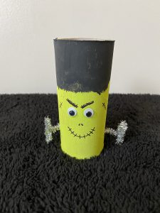 Trivallis Housing Landlord Wales A homemade craft resembling a Frankenstein figure. It is made from a painted toilet paper roll with a black top for hair, googly eyes, a drawn-on mouth and eyebrows, and metallic pipe cleaners for arms, set against a black textured surface.