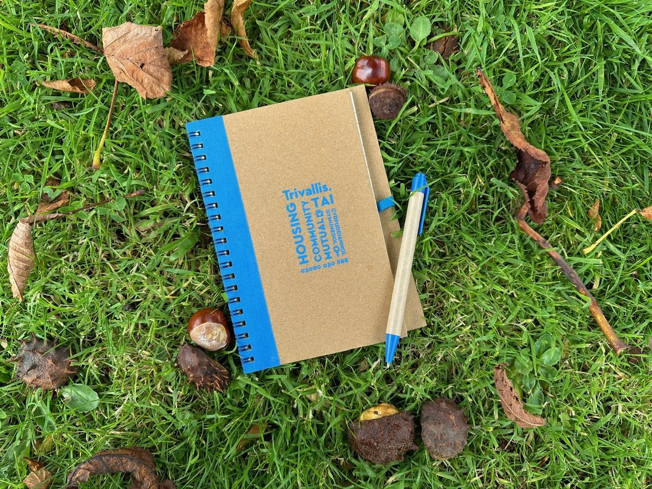 Trivallis Housing Landlord Wales An open spiral notebook with a cardboard cover and blue edge lies on green grass, surrounded by leaves, chestnuts, and a brown pen with a blue cap. The notebook cover reads "Housing, Community, Mutual Aid" in blue text.