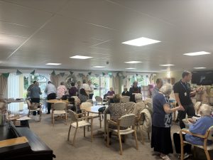 Trivallis Housing Landlord Wales A group of people gather in a community room with large windows, sitting at tables and standing while engaging in conversation. The room has chairs, tables, and a piano, with natural light coming in from outside.