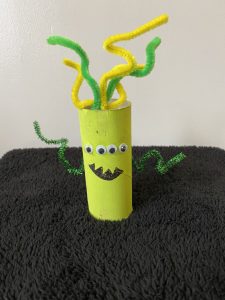 Trivallis Housing Landlord Wales A handmade craft made from a painted yellow toilet paper roll with attached googly eyes and a drawn mouth. Green and yellow pipe cleaners protrude from the top and sides, resembling arms and hair. The craft is placed on a black textured surface.