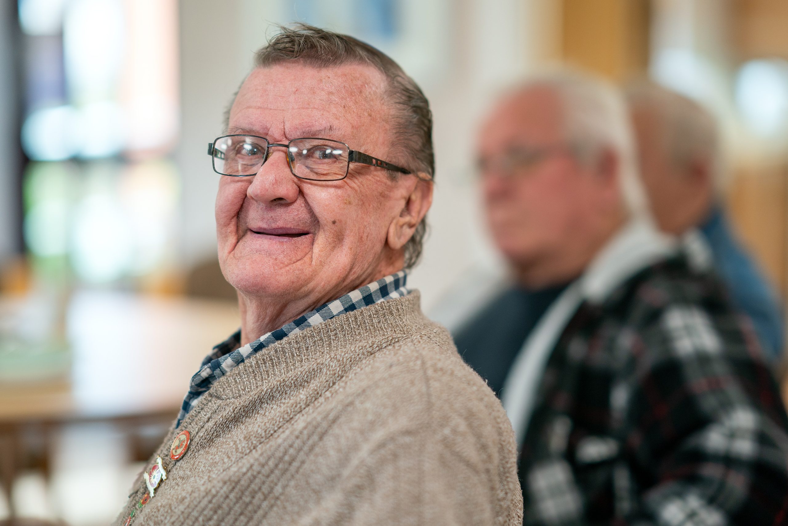 Trivallis Housing Landlord Wales An elderly man wearing glasses and a beige sweater is smiling while sitting in a room. He appears relaxed. In the background, another person is slightly out of focus, sitting in a plaid shirt. The setting seems casual and indoor.