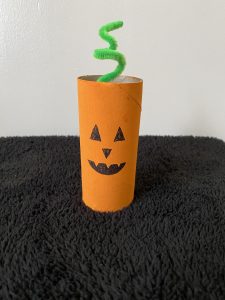 Trivallis Housing Landlord Wales A cardboard tube painted orange with a jack-o'-lantern face is standing on a black surface. A green pipe cleaner is inserted into the top, resembling a pumpkin stem.