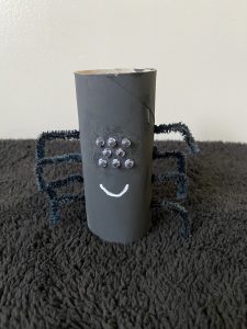 Trivallis Housing Landlord Wales A spider craft made from a painted toilet paper roll with googly eyes and black pipe cleaner legs. The roll is painted black with a white smile, and it rests on a dark, textured surface.