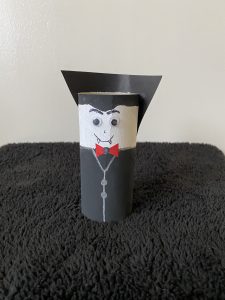 Trivallis Housing Landlord Wales A craft vampire made from a painted cardboard tube stands on a black textured surface. It features googly eyes, a red bow tie, and a black paper cape.