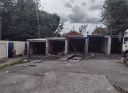 Trivallis Housing Landlord Wales A small, empty parking area with several open garages in a row. The ground is paved, and there is a truck partially visible on the right. Trees and a cloudy sky are in the background. Debris is scattered across the lot.