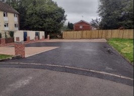 Trivallis Housing Landlord Wales A freshly paved driveway leads into an empty parking area, bordered by a wooden fence and flanked by trees. Brick pillars are present on the left, and a residential building is partially visible in the background.