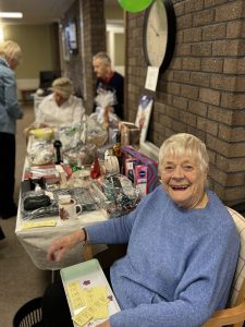 Trivallis Housing Landlord Wales An elderly woman in a blue sweater sits smiling at a table displaying various raffle prizes. Behind her, several people stand near the table in a community room. A wall clock and brick wall are visible in the background.