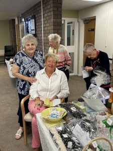 Trivallis Housing Landlord Wales Four women are in a room with a table displaying items for a raffle. One woman is seated, holding tickets, while the others stand nearby. A brick wall and a door are in the background. The table holds various items and raffle tickets.