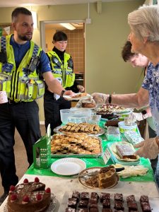 Trivallis Housing Landlord Wales A police officer in uniform receives a piece of cake from a person at a table filled with various pastries and cakes. Another officer and a few other individuals are in the background. The setting appears to be a community event.