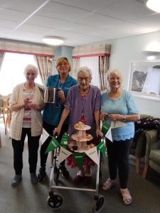 Trivallis Housing Landlord Wales Four women stand together indoors. One holds a coffee pot, another leans on a walker decorated with a small cake stand and festive flags. They appear to be in a communal living area with chairs and a painting on the wall.