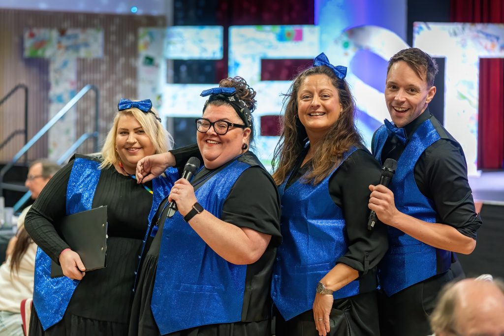 Trivallis Housing Landlord Wales A group of four people wearing black and blue outfits are standing together. Two of them hold microphones. They are smiling and posing indoors, with large, partially visible illuminated letters in the background.