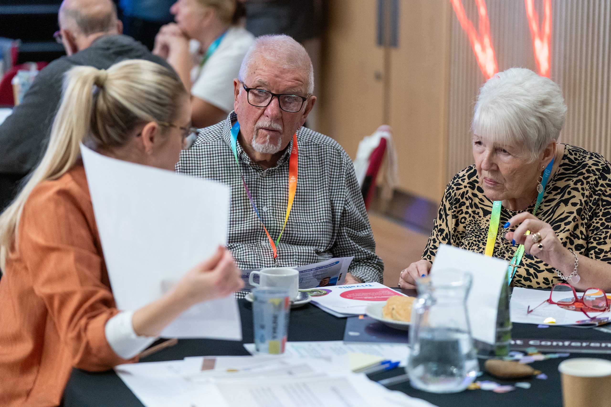 Trivallis Housing Landlord Wales A woman shows a document to two elderly people sitting at a table during a meeting. They are wearing conference lanyards. Papers, a mug, and a pitcher of water are on the table. Other attendees are visible in the background.