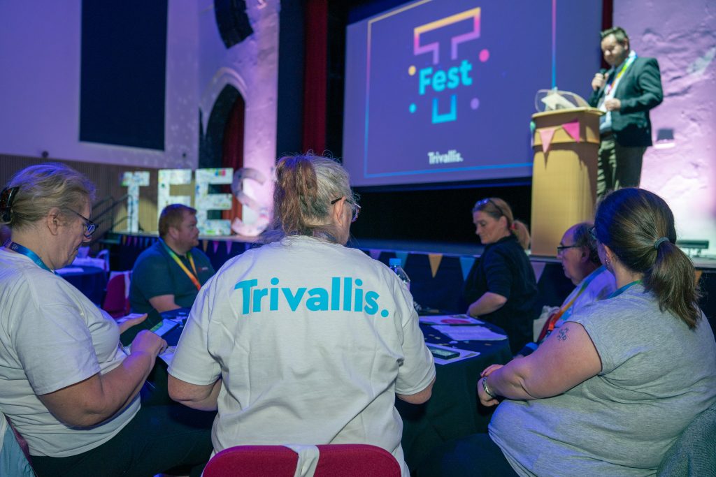 Trivallis Housing Landlord Wales A group of people sit around a table at an event. One person at a podium speaks to the audience, while a screen displays "T Fest" and the logo for Trivallis. Some attendees wear shirts with "Trivallis" printed on the back.