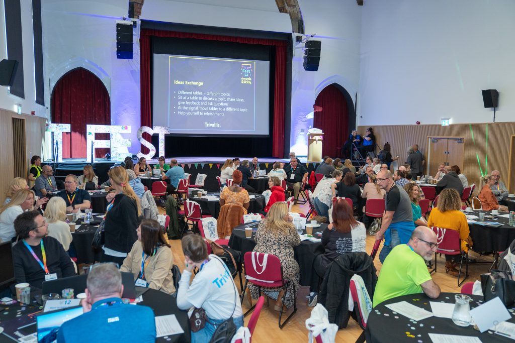 Trivallis Housing Landlord Wales A large group of people sit at circular tables in a conference hall, engaged in discussion. A screen displays "Ideas Exchange" with bullet points. A speaker stands at a podium near the stage. The room has high ceilings and arched windows.