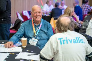 Trivallis Housing Landlord Wales Two people are seated at a table during an event. The person facing the camera is wearing a denim shirt and has event badges. The person with their back to the camera has "Trivallis" on their shirt. Papers and a coffee cup are on the table.