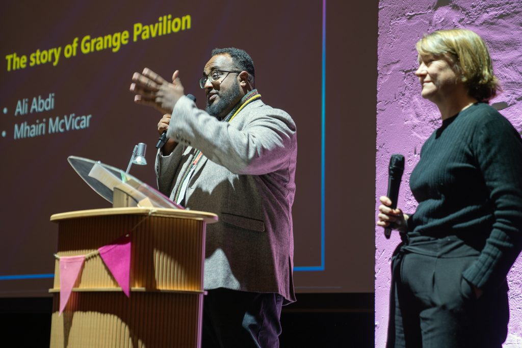 Trivallis Housing Landlord Wales A man and a woman are speaking on stage during a presentation. The man gestures while holding a microphone. Behind them is a screen displaying text about the story of Grange Pavilion, mentioning Ali Abdi and Mhairi McVicar.