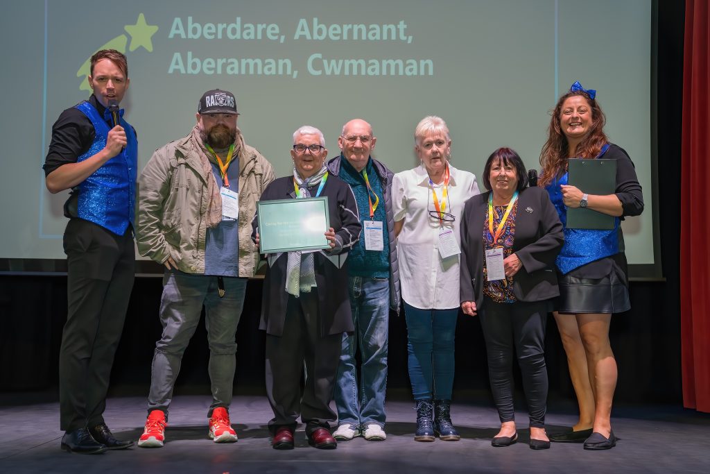 Trivallis Housing Landlord Wales A group of seven adults stand on a stage, posing for a photo. One person holds a framed certificate. Some are casually dressed, while two wear blue vests and another holds a microphone. A screen displays text about several towns in the background.