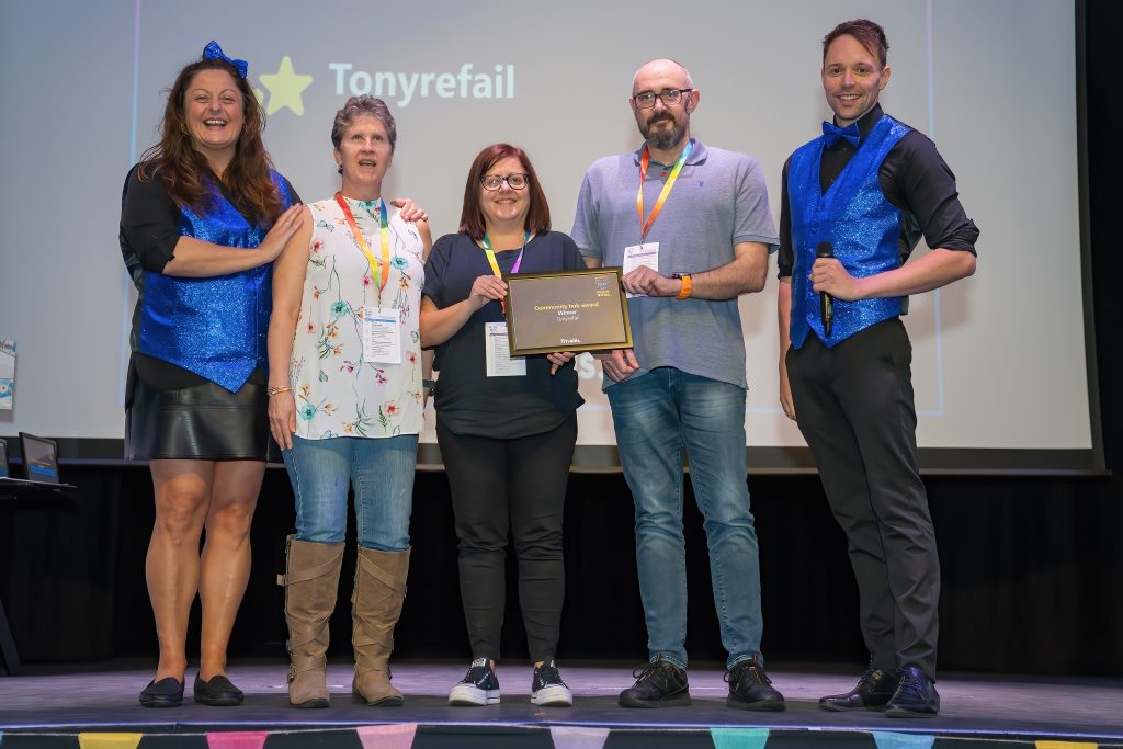 Trivallis Housing Landlord Wales Five people stand on stage. Three in the center hold a plaque. Two on the ends wear matching blue vests. A sign reading "Tonyrefail" is projected behind them. They appear to be celebrating or participating in an event.