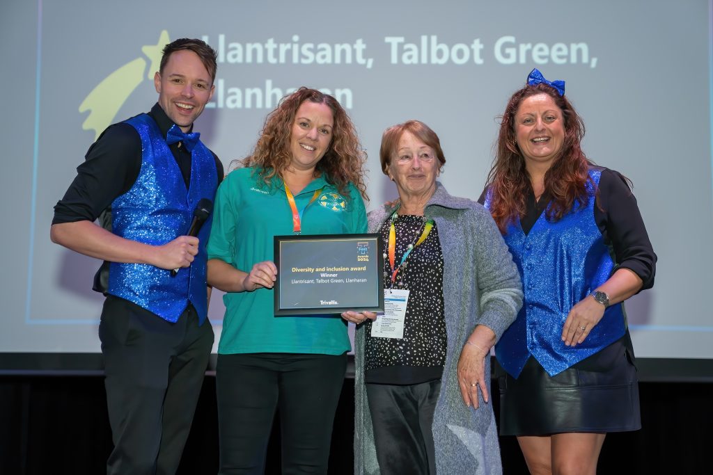 Trivallis Housing Landlord Wales Four people are standing on stage. Two are wearing sparkly blue outfits, one is holding a framed award, and another is in a gray cardigan. A large screen in the background displays text. They are smiling and posing for a photo.