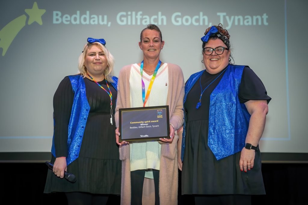 Trivallis Housing Landlord Wales Three people standing on stage, two wearing blue outfits, holding a framed award. The person in the middle is wearing a light-colored cardigan. A projected slide in the background reads "Beddau, Gilfach Goch, Tynant.